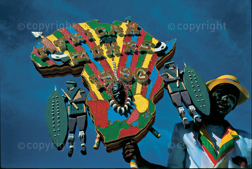  2 January 1996 - A member of the ‘African Zonks’ troupe brandishes his team’s bord at the Cape Minstrel Carnival ‘Annuals’. Since the inception of the new South Africa there have been attempts by organisers to make the carnival more culturally inclusive. This year a traditional Xhosa troupe was included in the programme for the first time in its history. The use of African symbols like the spear and shield on this bord indicate a move away from the use of imported symbols of the past, such as the Apache (North American Indian) themes characterised by feathered headgear and hatchets, so popular with troupes like the ‘Atchas’. President Nelson Mandela was the guest of honour on this occasion, making it a historic day for many. 
 The Kaapse Klopse (or simply Klopse), formerly known as the Coon Carnival and officially called Cape Town Minstrel Carnival, is a Cape coloured minstrel festival that takes place annually on 2 January in Cape Town, South Africa. It is also referred to as Tweede Nuwe jaar (Second New Year). As many as 13,000 minstrels take to the streets garbed in bright colours, either carrying colourful umbrellas or playing an array of musical instruments. The minstrels are self-organised into klopse (