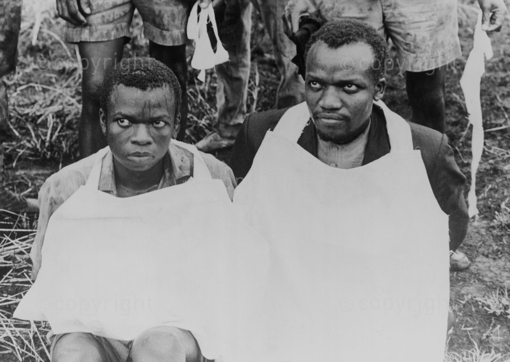 DM2008061805:EAED:POLITICS:PERSONALITIES:APR 1973 - Ugandans Flock To Public Executions - Fear shows clearly in the eyes of Sebastiano Namirundu (left) and Tom Masaba, two of the 12 alleged guerrillas executed.  They bound them to trees first.  Then they stripped them naked in front of 20,000 people.  They dressed them in aprons - white aprons, easy to see from a distance.  Then they shot them dead.  And as they cut them down in the pouring rain, the bleeding bodies fell into the mud.  Tom Masabe and Sebastiano Namirundu had seemed calm - or frozen with fear - before General Amin's soldiers formed a firing squad in that sodden-wet field at Mbale and carried out the public executions.  (Photograph by Drum Photographer ©BAHA)