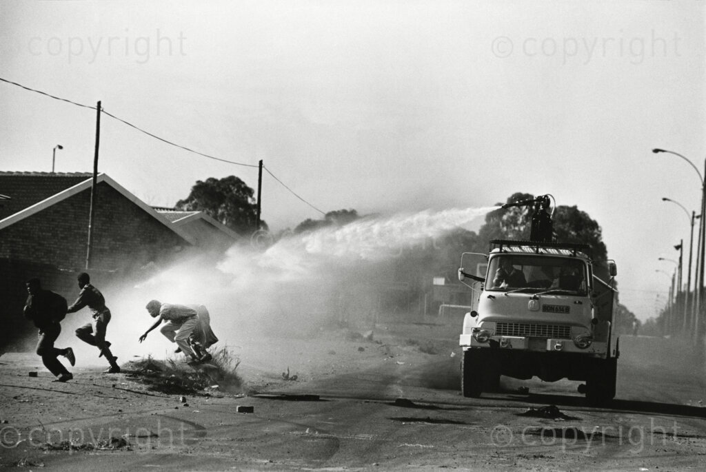 South Africa, 1990. Kagiso comrades get a dousing from a passing police fire-tender on its way to extinguish the burning municipal workshop - torched by other angry 'comrades'.
Kagiso is a township situated in the Krugersdorp area west of Johannesburg. The word 'kagiso' means 'peace' in Tswana.
Photo Credit: Ken Oosterbroek / african.pictures Photo Credit: Ken Oosterbroek / african.pictures