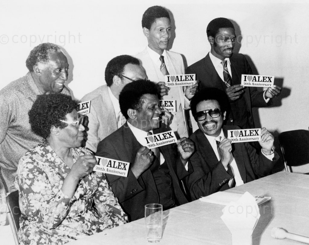October 1983. Alexandra Liaison Committee all holding stickers I love [heart] Alex 70th anniversary'. Front Row, from Left to Right: Mrs A. Serote, Rev. Sam Buti, Mr. Arthur Magerman. Back Row, from Left to Right: Mr. Loucas Khoza, Mrs. Percy Williams, Mr. Lewis Buti and Mr. Gerald Maseko. Photographer unknown. Â© Sunday Times / Times Media