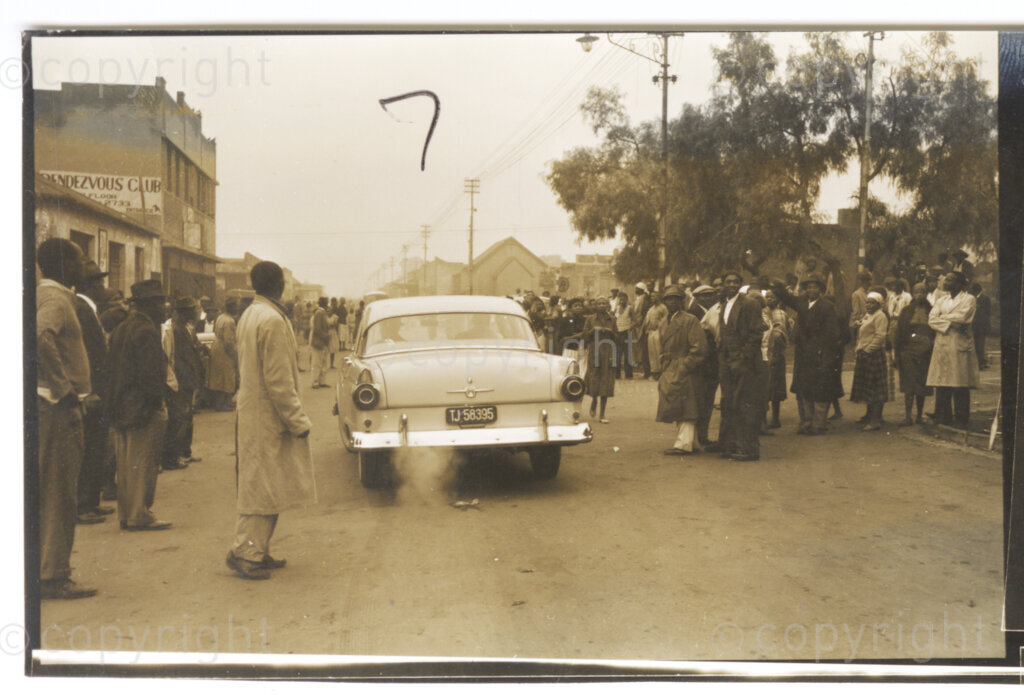 , Johannesburg, Transvaal (now Gauteng), South Africa, 19580414. PHOTO: Eli Weinberg.
A car drives down a road and a crowd of people mill around in a township somewhere near Johannesburg. A building with the name Rendezvous Club can be seen. This is part of a 'stay-at-home' called by the African National Congress (ANC) to protest the whites-only general election in South Africa on the 14-16 April 1958.