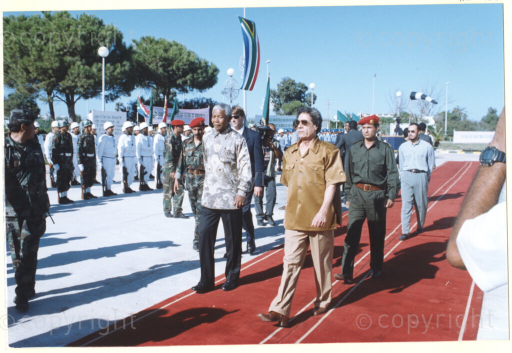 Tripoli, Tripoli District , Great Socialist People's Libyan Arab Jamahiriya (Libyan Arab Republic) , 19971022. PHOTO: ANC Archives.

Nelson Mandela (left) and his host Libyan leader Muammar Mohammed Abu Minyar Gaddafi (right) walk together along a red carpet upon Nelson Mandela's arrival in Tripoli, Libya (officially the Great Socialist People's Libyan Arab Jamahiriya (Libyan Arab Republic)) on 22 October 1997. The South African flag flies in the background. This is the second official visit as president of South Africa that Mandela has made to Libya, the first being in 1994, shortly after the first democratic elections. Libya has been an ally to the anti-apartheid movements since Colonel Muammar Gaddafi came to power in 1969, providing funding and support to the African National Congress (ANC). South Africa bestowed the Order of Good Hope, one of its highest honours, on Muammar Gaddafi in 1997.