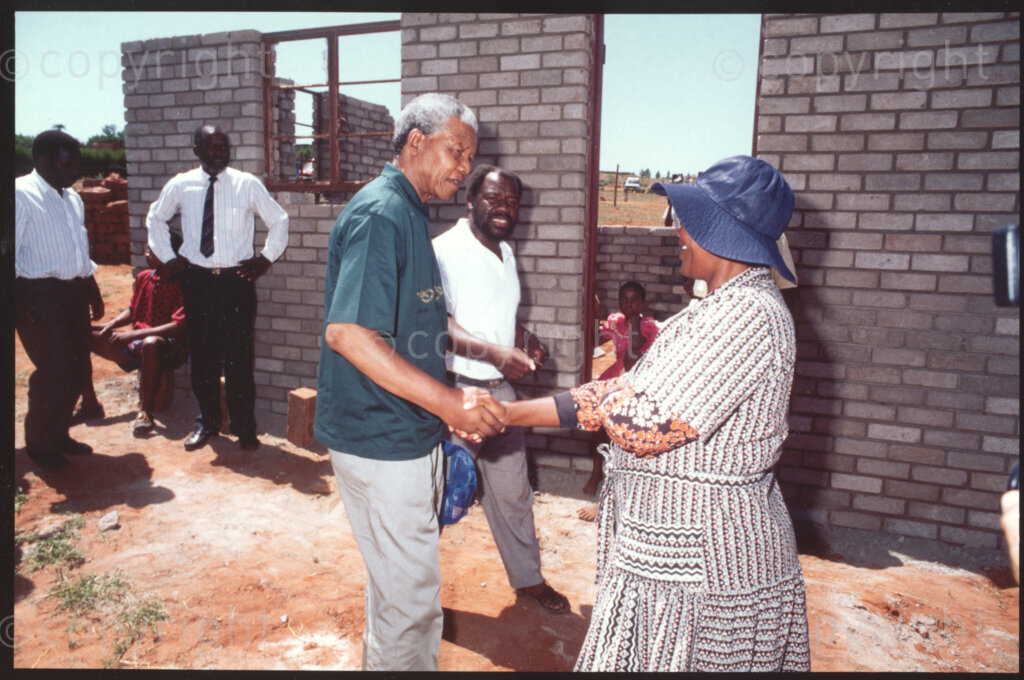 Kotsiri village, Sekhukhuneland, Transvaal, South Africa, 19921130. PHOTO: ANC Archives.

The President of the African National Congress, Nelson Mandela (centre), shakes hands with Elizabeth Shilakoe (right), an Operation Hunger field worker in Kotsiri Village, Sekhukhuneland in November 1992. The house behind them, which belongs to a family of orphan children, was destroyed during a recent storm and is being rebuilt by Operation Hunger.