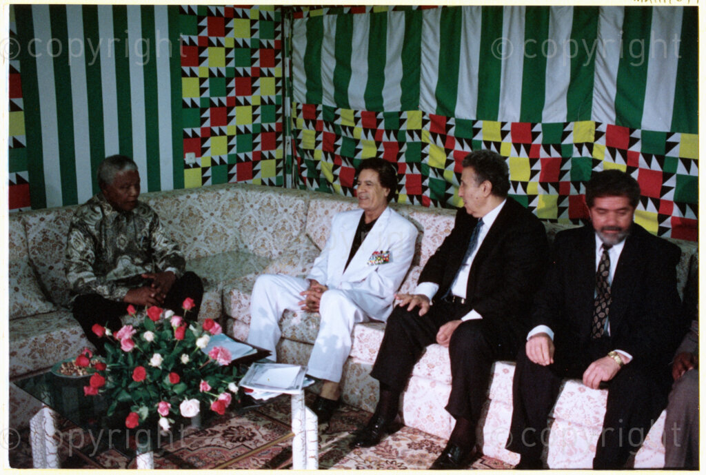 Tripoli, Tripoli District , Great Socialist People's Libyan Arab Jamahiriya (Libyan Arab Republic) , 19971022. PHOTO: ANC Archives.

Nelson Mandela (left), his host Libyan leader Muammar Mohammed Abu Minyar Gaddafi (second left), and two unknown Libyan officials sit together at a social function in Tripoli, Libya (officially the Great Socialist People's Libyan Arab Jamahiriya (Libyan Arab Republic)) on 22 October 1997. This is the second official visit as president of South Africa that Mandela has made to Libya, the first being in 1994, shortly after the first democratic elections. Libya has been an ally to the anti-apartheid movements since Colonel Muammar Gaddafi came to power in 1969, providing funding and support to the African National Congress (ANC). South Africa bestowed the Order of Good Hope, one of its highest honours, on Gaddafi in 1997.