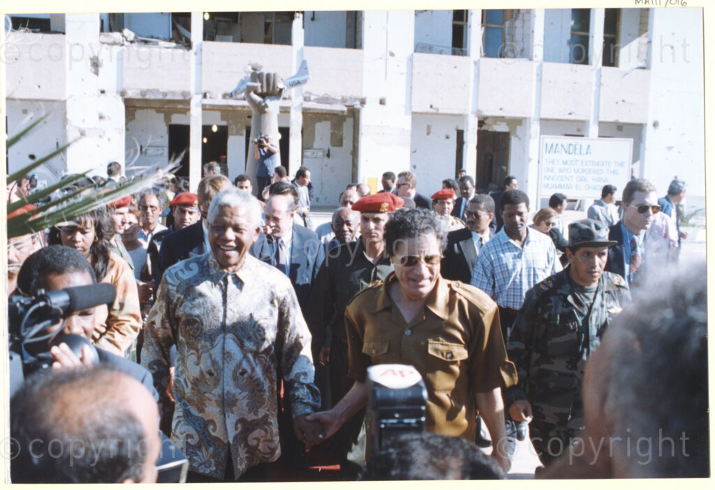 Tripoli, Tripoli District , Great Socialist People's Libyan Arab Jamahiriya (Libyan Arab Republic) , 19971022. PHOTO: ANC Archives.

Nelson Mandela (left) and his host Libyan leader Muammar Mohammed Abu Minyar Gaddafi (right) walk through a crowd (of members of the press and the public) that has assembled in front of a building destroyed by a U.S. fighter strike a decade earlier in Tripoli, Libya (officially the Great Socialist People's Libyan Arab Jamahiriya (Libyan Arab Republic)) on 22 October 1997. This is the second official visit as president of South Africa that Mandela has made to Libya, the first being in 1994, shortly after the first democratic elections. Libya has been an ally to the anti-apartheid movements since Colonel Muammar Gaddafi came to power in 1969, providing funding and support to the African National Congress (ANC). South Africa bestowed the Order of Good Hope, one of its highest honours, on Gaddafi in 1997.
