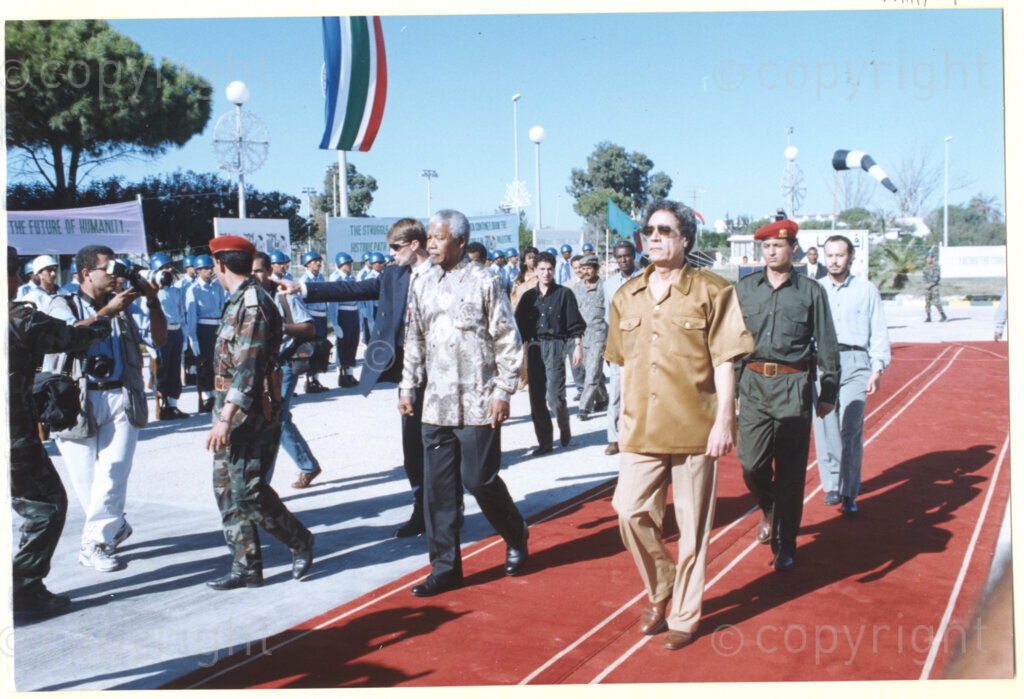 Tripoli, Tripoli District , Great Socialist People's Libyan Arab Jamahiriya (Libyan Arab Republic) , 19971022. PHOTO: ANC Archives.

Nelson Mandela (left) and his host Libyan leader Muammar Mohammed Abu Minyar Gaddafi (right) walk together along a red carpet upon Nelson Mandela's arrival in Tripoli, Libya (officially the Great Socialist People's Libyan Arab Jamahiriya (Libyan Arab Republic)) on 22 October 1997. The South African flag flies in the background. This is the second official visit as president of South Africa that Mandela has made to Libya, the first being in 1994, shortly after the first democratic elections. Libya has been an ally to the anti-apartheid movements since Colonel Muammar Gaddafi came to power in 1969, providing funding and support to the African National Congress (ANC). South Africa bestowed the Order of Good Hope, one of its highest honours, on Gaddafi in 1997.