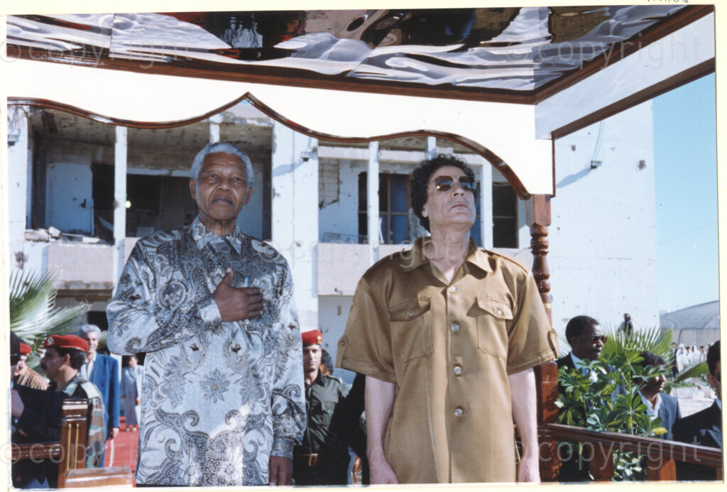 Tripoli, Tripoli District , Great Socialist People's Libyan Arab Jamahiriya (Libyan Arab Republic) , 19971022. PHOTO: ANC Archives.

Nelson Mandela (left) stands with his right arm folded across his chest next to his host Libyan leader Muammar Mohammed Abu Minyar Gaddafi (right) under an awning in front of building destroyed by a U.S. fighter strike a decade earlier, in Tripoli, Libya (officially the Great Socialist People's Libyan Arab Jamahiriya (Libyan Arab Republic)) on 22 October 1997. This is the second official visit as president of South Africa that Mandela has made to Libya, the first being in 1994, shortly after the first democratic elections. Libya has been an ally to the anti-apartheid movements since Colonel Muammar Gaddafi came to power in 1969, providing funding and support to the African National Congress (ANC). South Africa bestowed the Order of Good Hope, one of its highest honours, on Gaddafi in 1997.