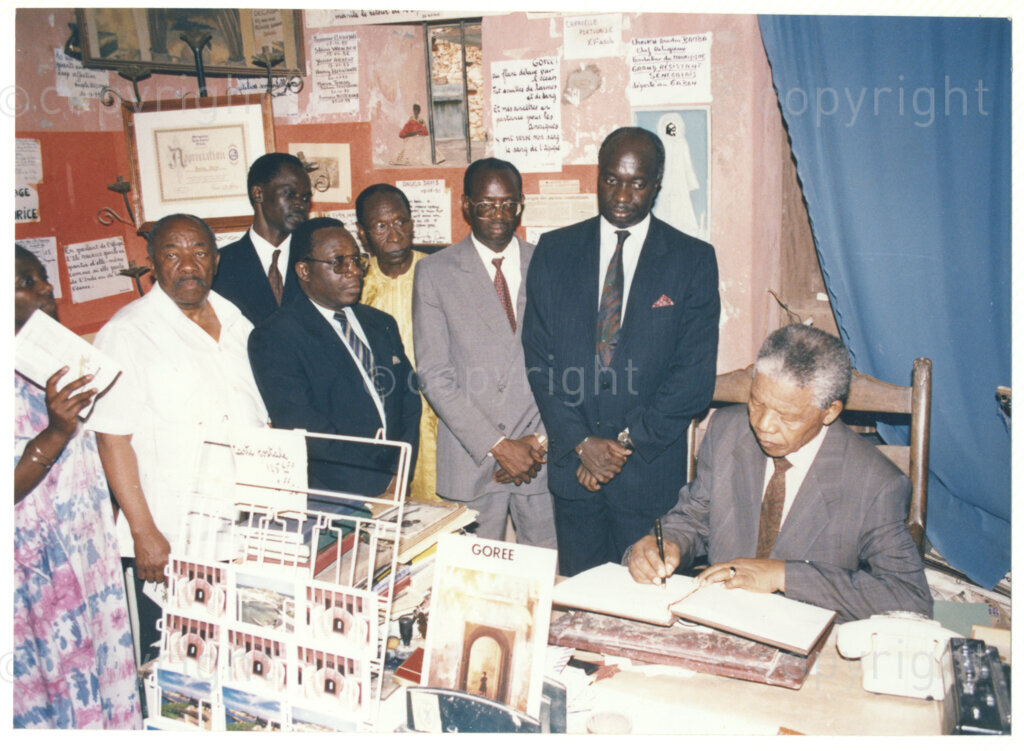 Senegal, 19920601. PHOTO: ANC Archives.

A group photo with Alfred Nzo (second from left) and Nelson Mandela (right) sitting at a table, probably signing a register, possibly on Goree Island, off Dakar, previously a large slave-trading centre, during his visit to Senegal in June 1992.