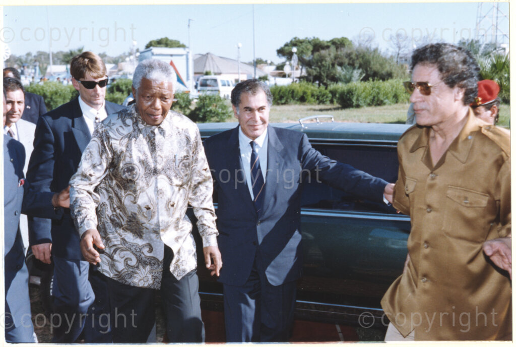 Tripoli, Tripoli District , Great Socialist People's Libyan Arab Jamahiriya (Libyan Arab Republic) , 19971022. PHOTO: ANC Archives.

Nelson Mandela (left, attended by one of his bodyguards) is ushered by an official towards his host Libyan leader Muammar Mohammed Abu Minyar Gaddafi (foreground right) upon his arrival in Tripoli, Libya (officially the Great Socialist People's Libyan Arab Jamahiriya (Libyan Arab Republic)) on 22 October 1997. This is the second official visit as president of South Africa that Mandela has made to Libya, the first being in 1994, shortly after the first democratic elections. Libya has been an ally to the anti-apartheid movements since Colonel Muammar Gaddafi came to power in 1969, providing funding and support to the African National Congress (ANC). South Africa bestowed the Order of Good Hope, one of its highest honours, on Muammar Gaddafi in 1997.