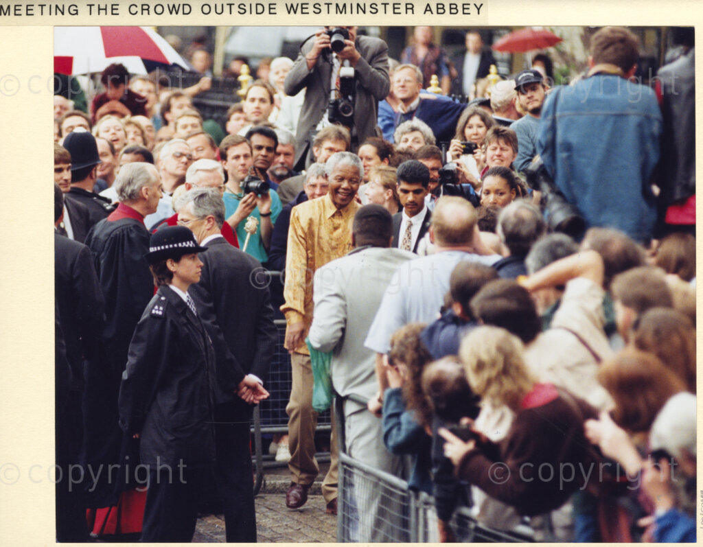 Outside Westminster Abbey, London, United Kingdom, 19960701. PHOTO: ANC Archives: During his July 1996 state visit to the United Kingdom, (UK), crowds of admirers crammed the streets of London, to get a glimpse of President Nelson Mandela, the iconic South African philanthropist. Mandela, touched by their fervour, is warmly responsive. Here he is seen, (wearing a gold shirt), outside Westminster Abbey.