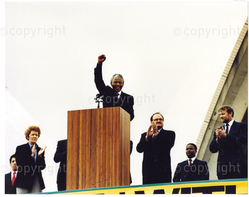 Australia, October 1990. PHOTO: ANC Archives: Nelson Mandela, Deputy President of the African National Congress, raises a fist in a solidarity salute after an address to his Australian audience, in 1990. He receives a standing ovation and is flanked by dignitaries and politicians at the Sydney Opera House. Monday 22 October to Saturday 27 October 1990.