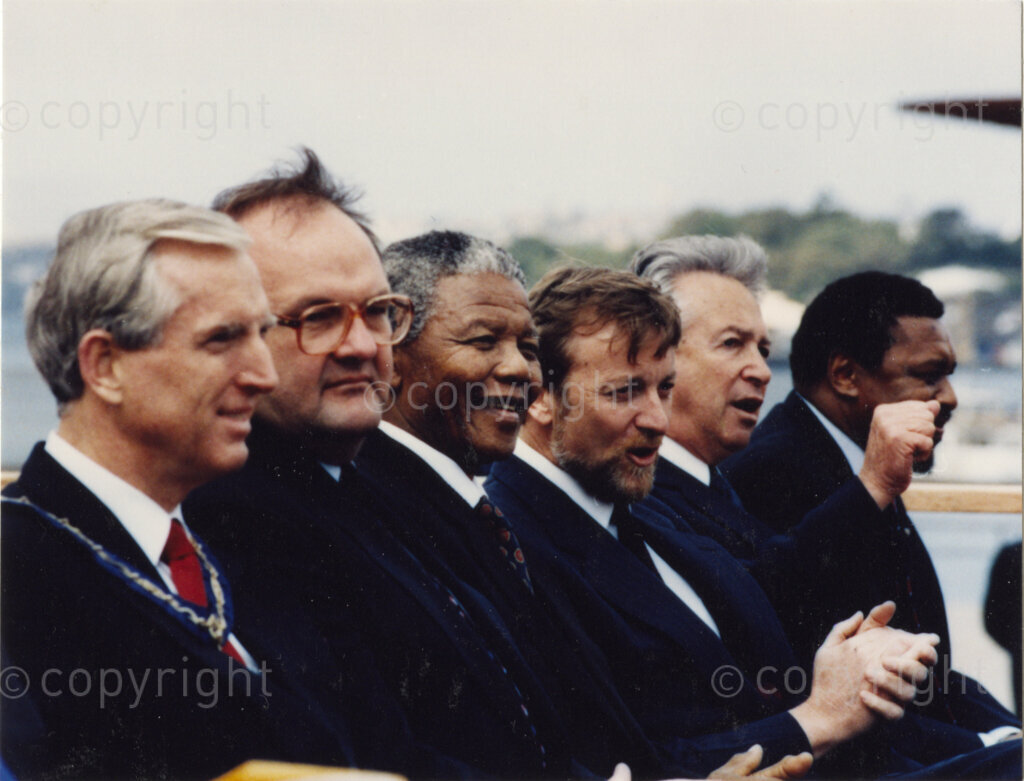 Australia, October 1990. PHOTO: ANC Archives: Nelson Mandela, during his 1990 visit to Australia, is a member of an appreicative audience that gives a round of applause to the speaker. Mandela, himself, addresses audiences throughout his visit. His mission is to thank Australia for their support, and to urge them to maintain the pressure on the oppressive apartheid regime. (Mandela is third from the left in the image. The other men are unnamed.) Monday 22 October to Saturday 27 October 1990.