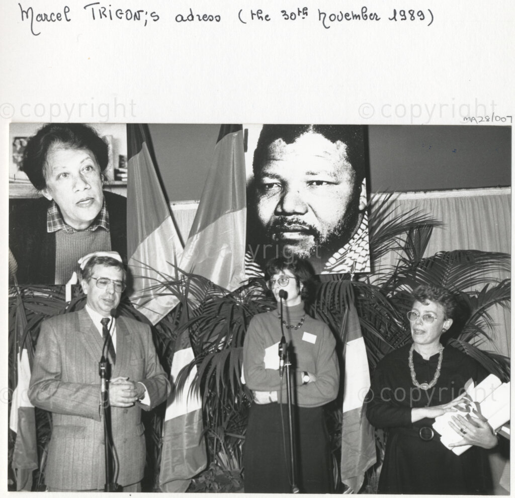 Arcueil, Val-de-Marne, Paris, Île-de-France, France, 19891130. PHOTO: ANC Archives: Mayor of Arcueil (Val-de-Marne) and figure of communism, Marcel Trigon (left) addresses a crowd of people at a function in Arcueil, France on 30 November 1989. A number of representatives from a variety of French anti-apartheid groups and African National Congress comrades are attending the gathering to commemorate Dulcie September and Nelson Mandela, images of whom appear in large posters behind the podium. Jacqueline Derens, general secretary of National Gathering against Apartheid (RNCA) (right) looks on. Dulcie September, a South African anti-apartheid activist and ANC Chief Representative in France, Switzerland and Luxembourg, was assassinated outside the ANC's Paris office at 28 Rue des Petites-Ecuries last year on 29 March 1988.