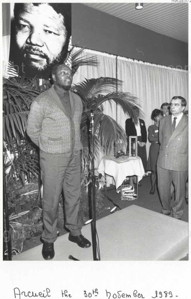 Arcueil, Val-de-Marne, Paris, Île-de-France, France, 19891130. PHOTO: ANC Archives: An African National Congress (ANC) comrade makes a speech at a gathering in Arcueil, France on 30 November 1989 for the unveiling of a commemorative plaque for Dulcie September. The Mayor of Arcueil (Val-de-Marne) and figure of communism, Marcel Trigon (right) looks on. Behind the speaker is a large poster of Nelson Mandela who is imprisoned in South Africa. Dulcie September, a South African anti-apartheid activist and ANC Chief Representative in France, Switzerland and Luxembourg, was assassinated outside the ANC's Paris office at 28 Rue des Petites-Ecuries last year on 29 March 1988.