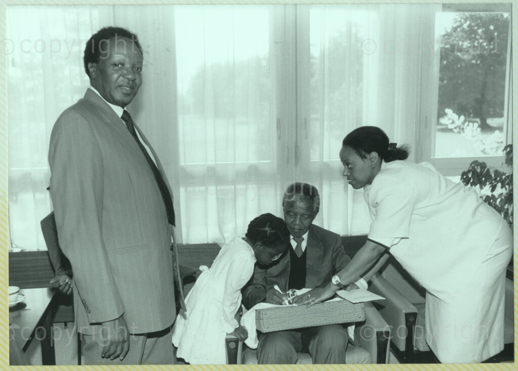 International Reformed Centre John Knox, Geneva, Switzerland, 19900609. PHOTO: ANC Archives.

Seated in a room with unidentified officials and a young girl Nehanda Mutambirwa, recently appointed Deputy President of the African National Congress (ANC) Nelson Mandela signs autographs at the International Reformed Centre John Knox (IRCJK), Geneva, Switzerland, 9 June 1990 during Nelson Mandela Mandela's official visit to Switzerland, 4-9 June 1990.