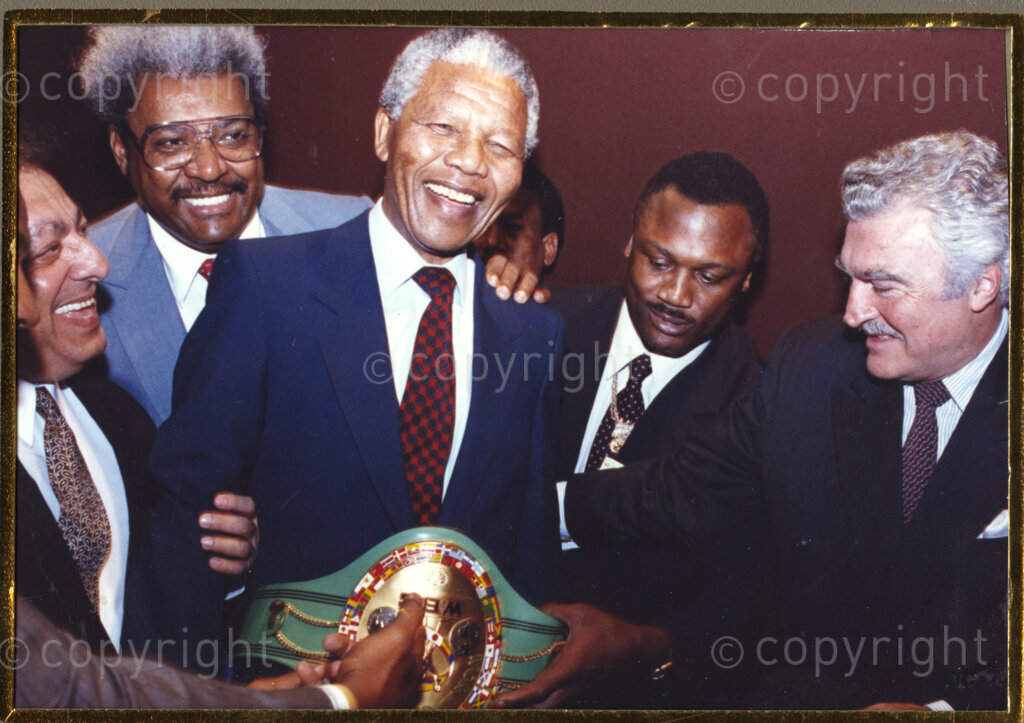 New York, New York State, United States of America, 19900601. PHOTO: ANC Archives.

In 1990, Nelson Mandela and his wife, Winnie Mandela, visit the city of New York from 20 to 23 June. This is the start of a 14-nations Freedom Tour, just 4 months after Mandela's release from jail. Wearing a boyish grin, Mandela, (centre), dons a boxing champion's belt. Peering over Mandela's shoulder is boxing promoter, Don King. Perhaps Mandela is reflecting on his own youthful boxing pursuits.