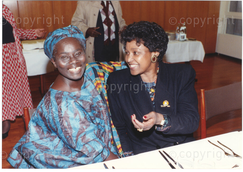 Geneva Ecumenical Centre, Geneva, Switzerland, 19900601. PHOTO: ANC Archives.

Winnie Madikizela-Mandela (right) and Ghanaian Methodist theologian Mercy Amba Oduyoye seated at a table smile broadly at the time of Nelson Mandela's visit to staff and officials and representatives of the World Council of Churches (WCC) at the Geneva Ecumenical Centre, 4-9 June 1990. During this visit, Nelson Mandela expresses his gratitude to the church for its support of the anti-apartheid struggle.