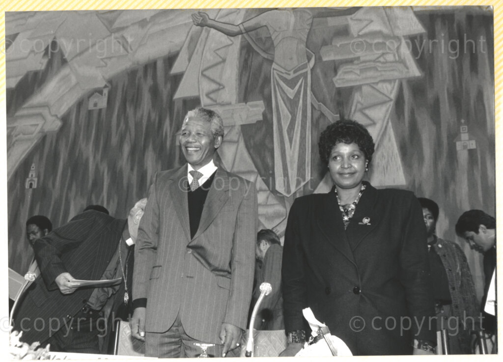 Geneva Ecumenical Centre, Geneva, Switzerland, 19900601. PHOTO: ANC Archives.

Standing behind the main table on stage recently elected Deputy President of the African National Congress (ANC) Nelson Mandela (left) and Winnie Madikizela-Mandela smile broadly amongst unidentified officials at the Geneva Ecumenical Centre, Switzerland when the Mandela couple visit staff, officials and representatives of the World Council of Churches (WCC) at the time of Nelson Mandela's official visit to Switzerland, 4-9 June 1990. During the visit, barely a few months after his release from prison, Nelson Mandela expresses his gratitude to the church for its support of the anti-apartheid struggle