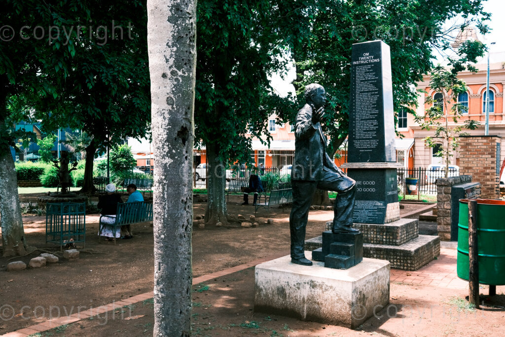 Figures in a 'Scape, Untitled sculpture of a gesturing adult man in a suit, located in a public space alongside the city hall, benches, Om plinth, rubbish bin and palm tree. The sculpture is just off Chief Albert Luthuli Road that runs through the center of this historic city and the city hall.