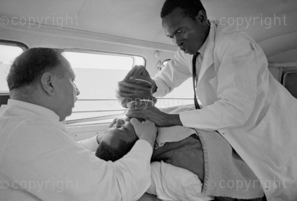 After Tom Mboya was shot outside the Pharmacy on July 5, 1969, the scene quickly turned chaotic as bystanders realized the gravity of the situation. Mboya, one of Kenya's most influential leaders, lay gravely wounded on the pavement. Within minutes, an ambulance arrived, and medical personnel, along with Dr. Chaudry, who had quickly come to the scene, worked urgently to get him onto a stretcher. Inside the ambulance, frantic efforts continued to try and save Tom Mboya.