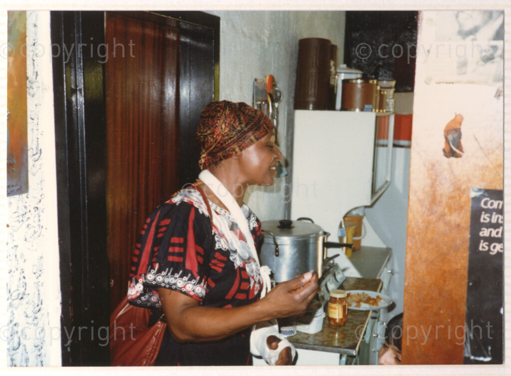 A portrait of Winnie Mandela standing in the doorway of what is thought to be her kitchen in her home in Brandfort, circa 1980. Winnie's left arm is in a sling and plaster cast, and she is wearing a red and black printed dress with white lace detail around the neck and a head scarf. Winnie was one of the most important icons of the struggle against apartheid in South Africa, and was forced to live in domestic exile in Brandfort, a small Afrikaner town in the Free State, for more than eight years.