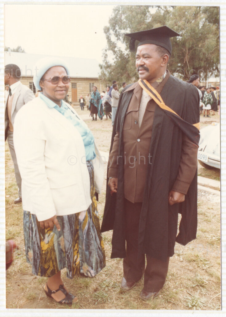 An unidentified man and woman stand next to each other during an event circa 1980. The man (right) is wearing a graduation gown, sash and mortarboard, and other people in the background are also wearing graduation outfits. This image is included in an album containing photographs from Winnie Mandela's time in Brandfort.