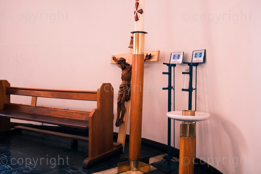Figures in a 'Scape. Crucified Christ. located behind an extra pew, candle holders and sanitary notification in the main area of the Cathedral of the Holy Nativity. The cathedral is the episcopal seat of the Bishop of Natal. The cathedral is located on Langalibalele Street. The cathedral was consecrated on 22 November 1981 and was designed by the architect firm of Kammeÿer, Rozendal and Carter-Brown. Oct. 2024, Pietermaritzburg, South Africa.
