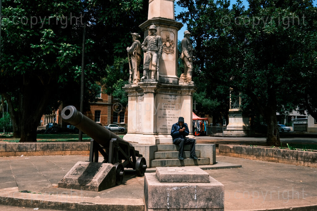 Figures in a 'Scape. A memorial to the Natal colonists who fall in the Anglo Zulu wars. A man smokes cigarette beneath the column in a park on the corner of Church street and Chief Albert Luthuli Road. Oct. 2024, Pietermaritzburg, South Africa.