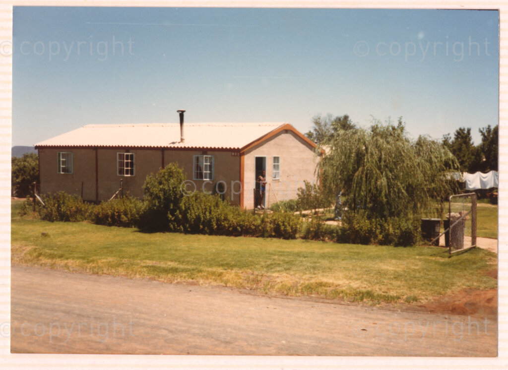 Winnie Mandela's house in Brandfort, 'House Number 802'. An unidentified person stands in the doorway of the small house, circa 1980. The simple building and garden is surrounded by a fence, and the property is next to a gravel road. White laundry is drying on a washing line at the back of the garden. Winnie was one of the most important icons of the struggle against apartheid in South Africa, and was forced to live in domestic exile in Brandfort, a small Afrikaner town in the Free State, for more than eight years.