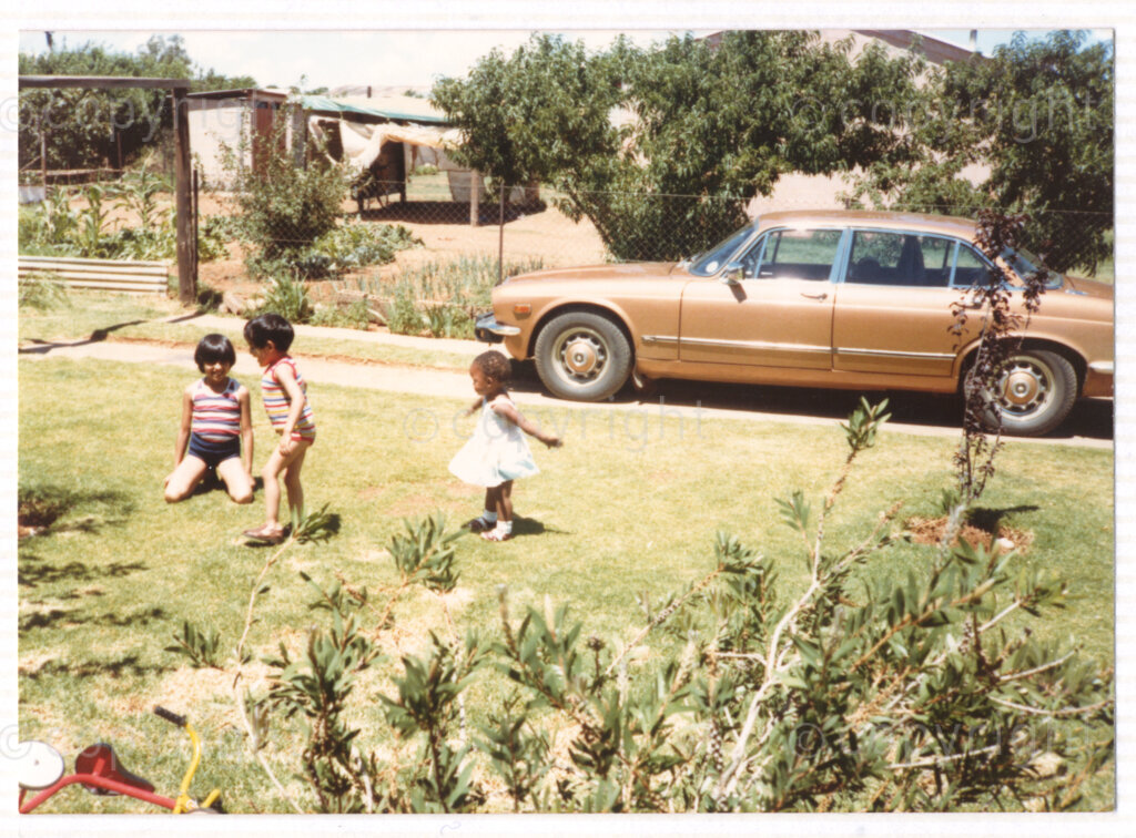 Three unidentified children looking relaxed and playful in a garden environment. The two older children (left) are white, and the younger girl (right) is black. The garden is thought to be Winnie Mandela's garden in Brandfort, and there is a brown car parked in the driveway. 
Winnie was one of the most important icons of the struggle against apartheid in South Africa, and was forced to live in domestic exile in Brandfort, a small Afrikaner town in the Free State, for more than eight years.