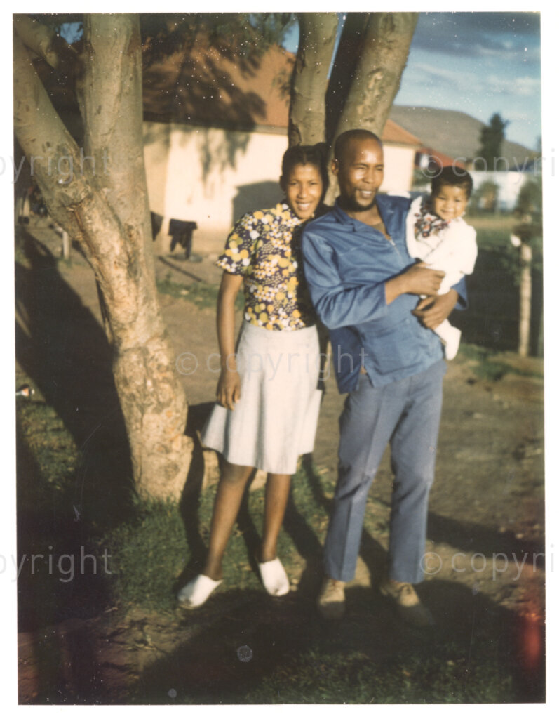 Three unidentified people (two adults, one baby) pose for a photograph beneath a tree. They appear to be a family and look happy and relaxed. This image is included in an album containing photographs from Winnie Mandela's time in Brandfort.