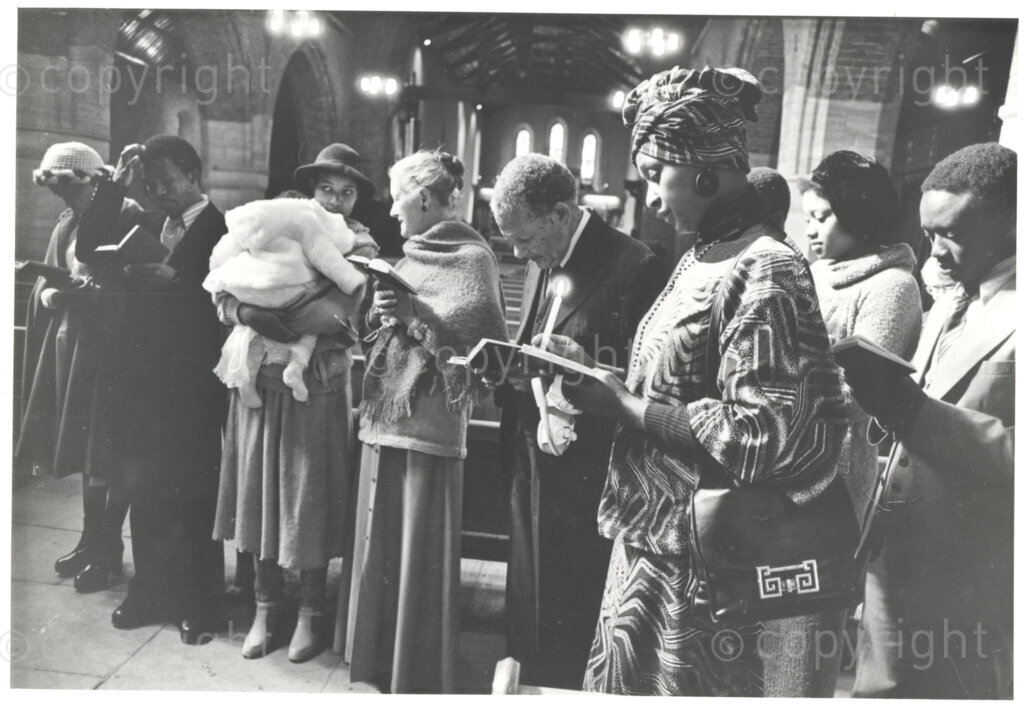 Winnie Mandela (right, front) stands in what is thought to be a church service, reading from a small hymn book, circa 1980. She is one of ten people in the church, ranging from a very young baby to an old man. Winnie is formally dressed in a printed outfit and matching headwear.
