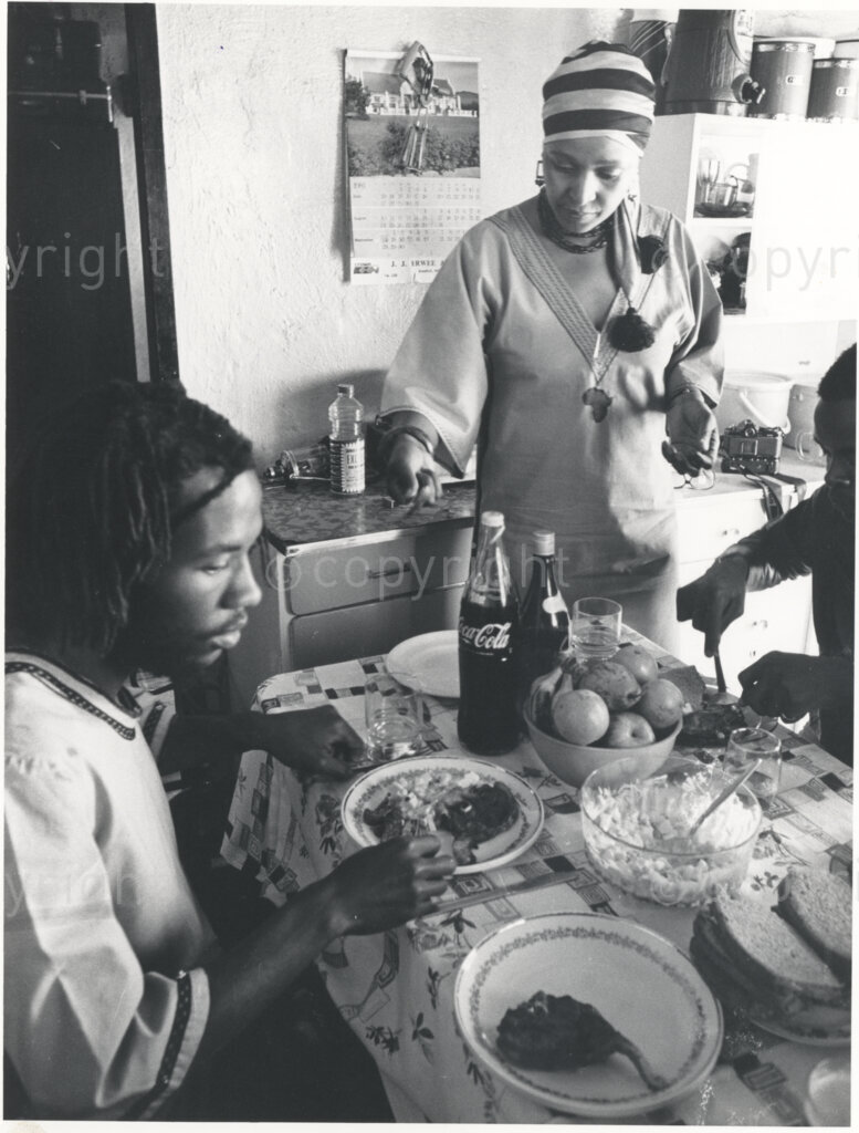 Winnie Mandela (centre) stands between two unidentified people at a laden table, circa 1980. The picture is thought to have been taken in the kitchen of Winnie's home in Brandfort, where she was forced to live in domestic exile for more than eight years. Winnie wears a striped head scarf, and the table is laden with food and drinks.