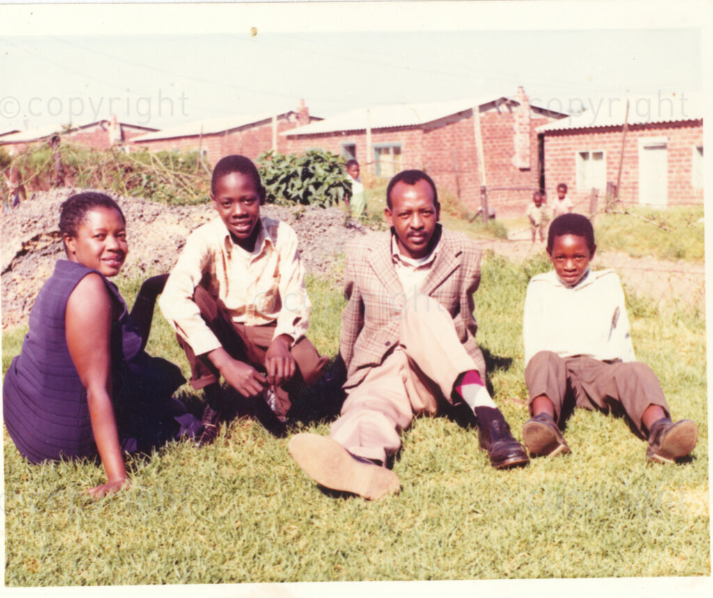 An unidentified woman (left), man (second from right) and two boys pose for a photograph, sitting on grass, circa 1980. What is thought to be the Brandfort township There are face-brick houses behind them, and three children in the background. The group is thought to be a family. This image is included in an album containing photographs from Winnie Mandelaâ€™s time in Brandfort. 