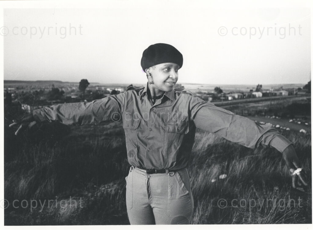 A portrait of Winnie Mandela wearing a military-style shirt and a dark beret, circa 1980. What is thought to be the town of Brandfort is visible in the distance. Winnie had endured imprisonment, torture and harassment for years before she was banished to this small Afrikaner town, hundreds of kilometers away from her home in Johannesburg. She lived in domestic exile in Brandfort for more than eight years.Â 