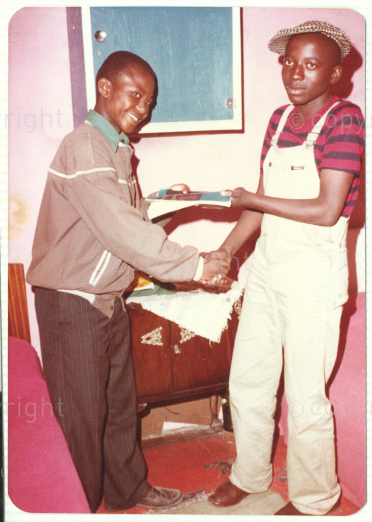Two unidentified teenage boys shake hands in what appears to be a ceremonial gesture, circa 1980. One (left) appears to be awarding the other (right) a certificate or a book. This image is included in an album containing photographs from Winnie Mandela's time in Brandfort.