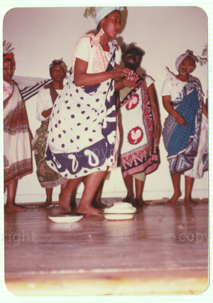 A group of five women in traditional dress on what is thought to be a stage, circa 1980. There are several white enamel bowls on the floor. This image is included in an album containing photographs from Winnie Mandela's time in Brandfort.