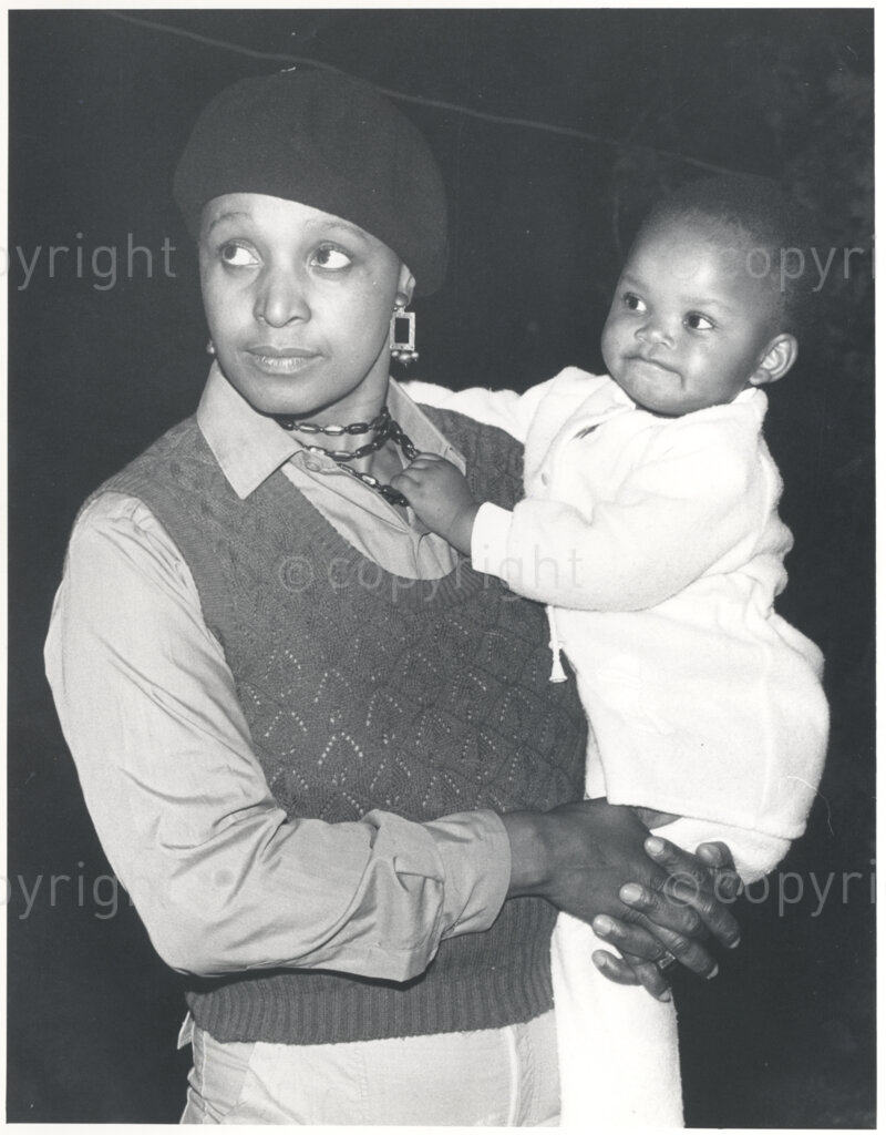 Winnie Mandela holds an unidentified baby, circa 1980. Winnie is wearing a dark beret and a shirt, and she and the baby are both looking at something or someone on their right (not pictured). Winnie was one of the most important icons of the struggle against apartheid in South Africa.