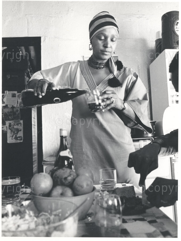 A portrait of Winnie Mandela pouring Coca-Cola into a glass, standing at a laden table in a kitchen circa 1980. She is wearing a scarf on her head. Mandela was one of the most important icons of the struggle against apartheid in South Africa, was forced to leave her home in Johannesburg under the Internal Security Act in 1977. She lived in domestic exile in the small town of Brandfort for more than eight years.