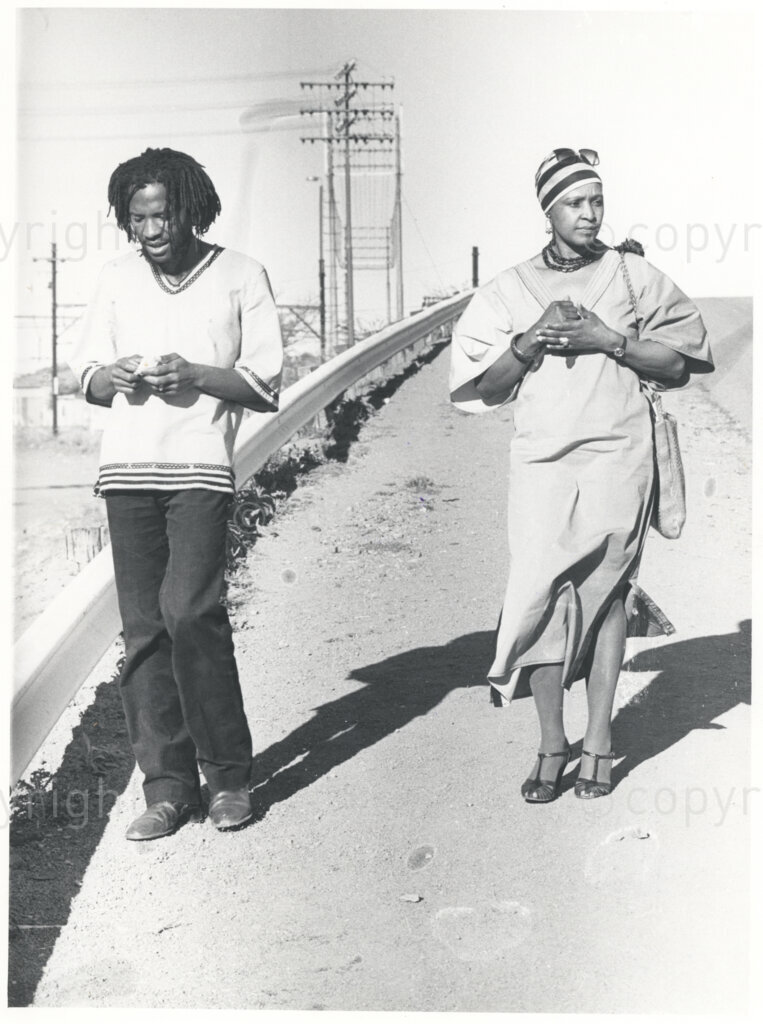 Winnie Mandela (right) and an unidentified man (left) walk down a dirt road, circa 1980. There are electricity poles behind them, and roadside crash barriers to their right. Winnie is dressed formally in a long dress, necklace and silk head scarf, and she is clasping her hands together.
Photo credit: ANC Archives / african.pictures