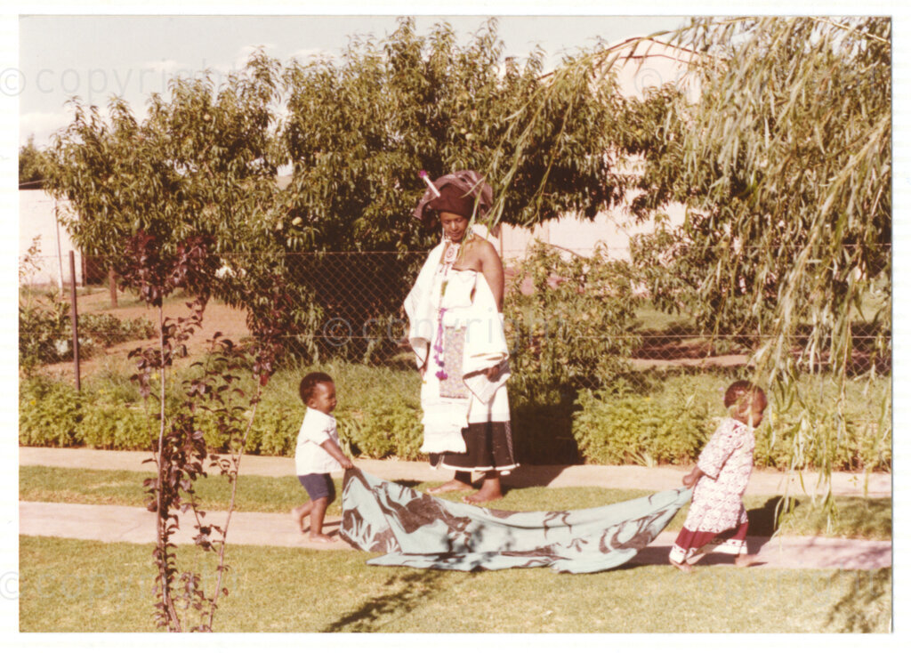 Winnie Mandela (centre) watching two unidentified young children and looking relaxed in a garden environment. The children are carrying what appears to be a blanket down a pathway. Winnie is wearing traditional Xhosa attire, and her predominantly cream and brown outfit and headwear reflects the cultural heritage of her birthplace, Bizana, a rural town in the Transkei in the Eastern Cape.Â Winnie was one of the most important icons of the struggle against apartheid in South Africa and wore traditional outfits and accessories during a time when donning cultural wear was seen as political taboo.