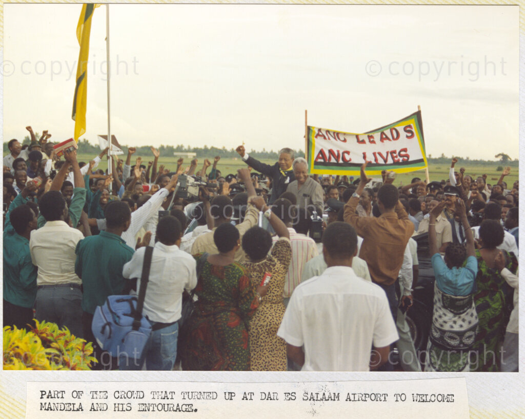 Dar Es Salaam, Tanzania. PHOTO: ANC Archives. 6 March 1990. An enormous crowd lined the route to the airport in Dar Es Salaam to welcome Nelson and Winnie Mandela. From the open top vehicle, Nelson Mandela (left) stands beside Julius Nyerera and waves to the crowds. A banner reads, 