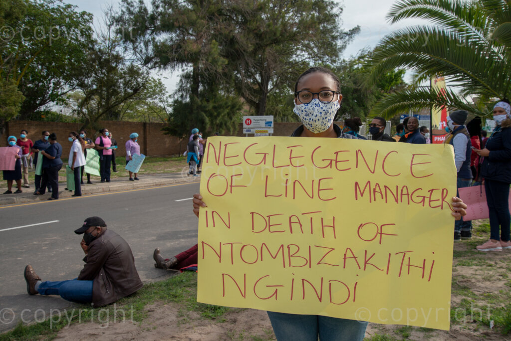 “I tested positive,” says Jemimah Mokubunge, on Friday, June 5, 2020, holding a (health-union SAFTU) sign that alleges that Tygerberg nurse Ntombizakithi Ngidi, who died of COVID-19 related illness, was initially refused medical care by her superiors. 
Mokubunge is a clerk handling patients’ hospital records. “Sometimes we get coronavirus patients’ records,” she said. “And there was no sanitizer. Most of the people (in the department) got positive.”
When the first person in her department contracted the virus, that employee was the one to notify her colleagues by calling them from isolation, explains Mokubunge. “We asked to be tested,” she said. But “they refused us to get tested.” 
“Only when the second person tested positive were we allowed to get tested,” she said. “I tested positive. I did not feel sick.” 
Mokubunge lives with her husband, mother and nine-month old baby. “They said I could isolate with the baby,” she said she was advised by the health professionals.  
After quarantining at home for 14 days, Mokubunge was told to go back to work. No retesting was available, she said. Her family tested negative for the virus. 
Hospital workers demonstrate outside Tygerberg Hospital, in Cape Town, to bring their plight to the attention of President Cyril Ramaphosa, as he visited the Cape Town Province. PHOTO: EVA-LOTTA JANSSON
